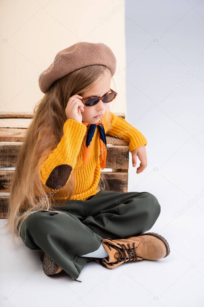fashionable blonde girl in autumn outfit and sunglasses posing near wooden box on beige and white background