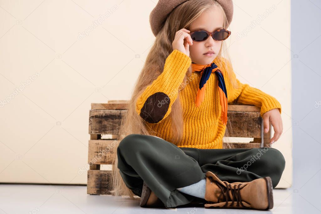 fashionable blonde girl in autumn outfit and sunglasses posing near wooden box on beige and white background