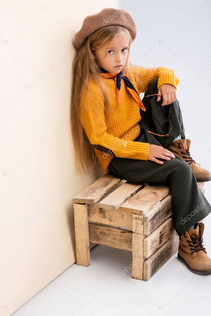 fashionable blonde girl in autumn outfit with sunglasses sitting on wooden box on beige and white background