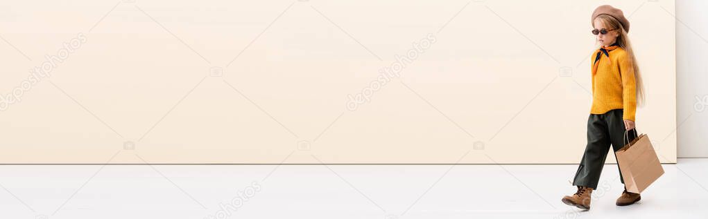 fashionable blonde girl in autumn outfit and sunglasses walking with shopping bag on beige and white background, panoramic shot