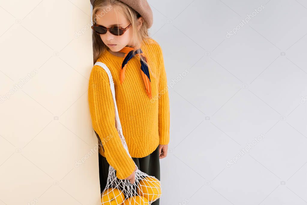 fashionable blonde girl in autumn outfit and sunglasses posing with grapefruits in string bag on beige and white background