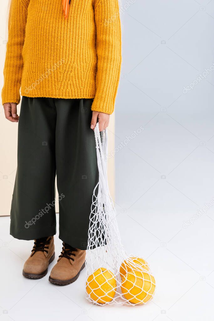 cropped view of fashionable girl in autumn outfit posing with grapefruits in string bag on beige and white background