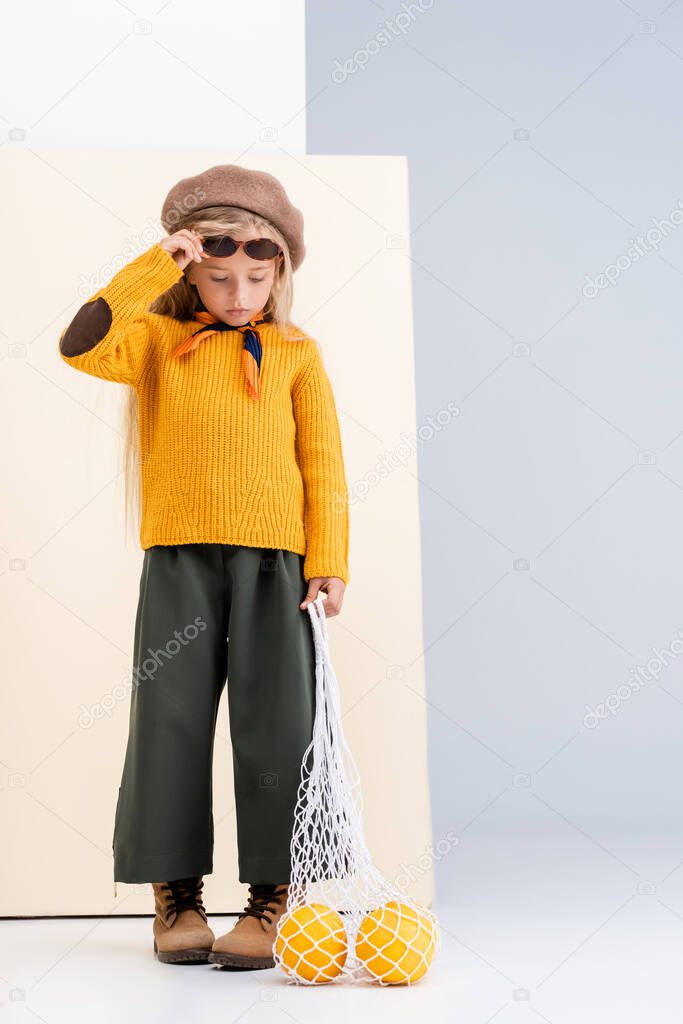 fashionable blonde girl in autumn outfit and sunglasses posing with grapefruits in string bag on beige and white background