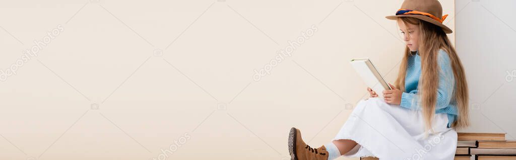 fashionable blonde girl in brown hat and boots, white skirt and blue sweater sitting on vintage books and reading near beige wall, panoramic shot