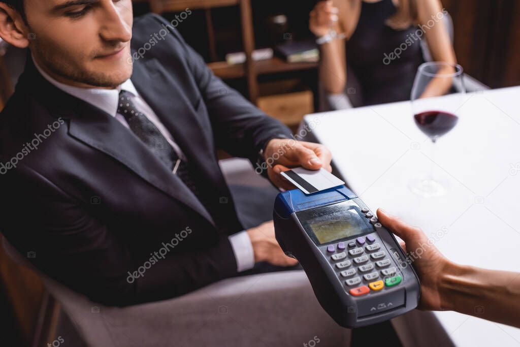 Selective focus of man in suit paying to waitress with payment terminal near girlfriend and glass of wine in restaurant 