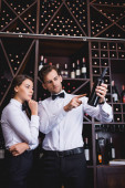 Sommelier pointing with finger ant bottle of wine near pensive colleague in restaurant 