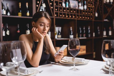 Restoranda bir kadeh şarabın yanında otururken akıllı telefon kullanan genç bir kadının seçici odağı. 