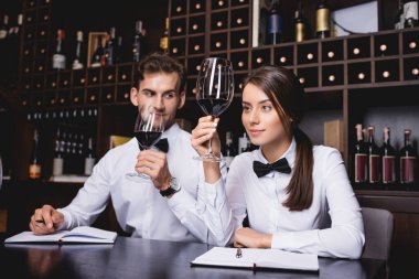 Selective focus of sommelier holding glass of wine near colleague and notebooks on table  clipart