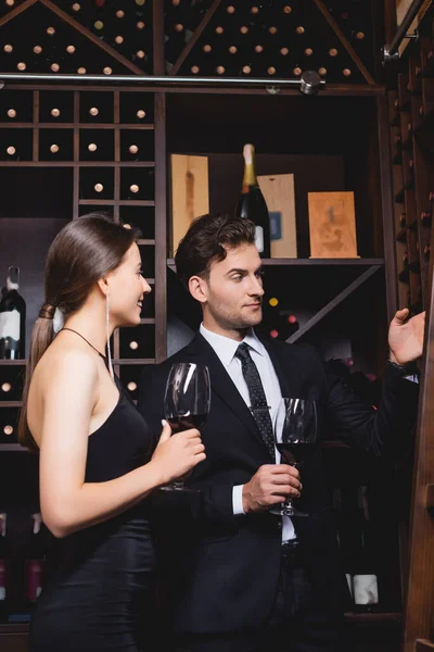 スーツ姿の男が手でラックで指差すエレガントな女性の近くのボトルレストランでワインのガラス — ストック写真