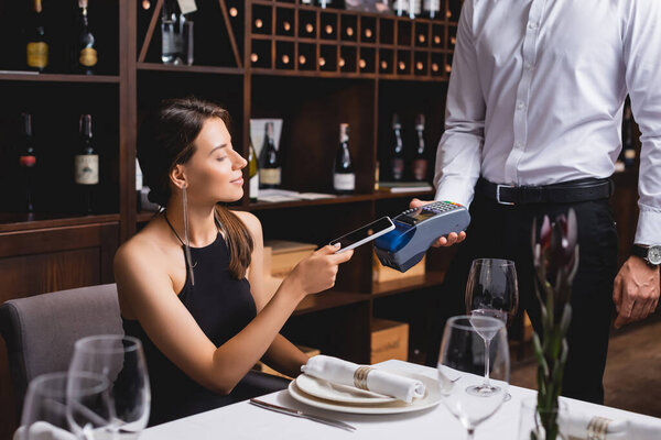 Selective focus of elegant woman paying with smartphone to waiter in restaurant 