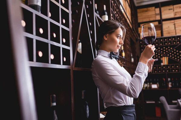 Selective focus of sommelier looking at wine in glass near racks with bottles 