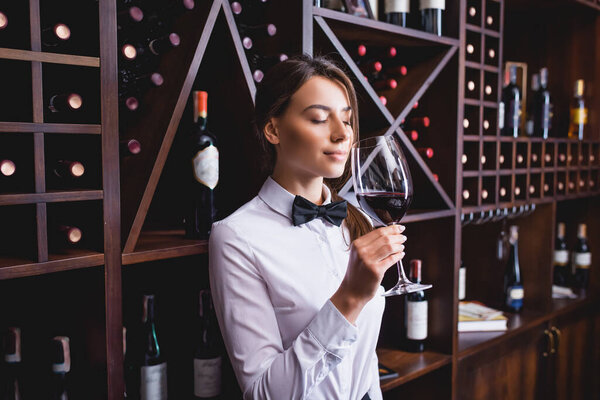 Young sommelier smelling glass of wine near racks with bottles 