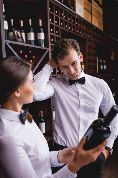 Selective focus of pensive sommelier with hand in pocket standing near colleague with bottle of wine 