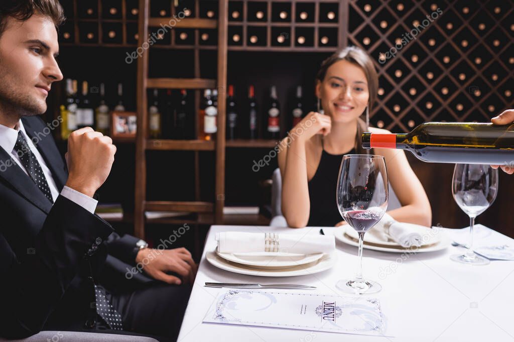 Selective focus of sommelier pouring wine in glass near elegant couple sitting at table in restaurant 