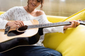 oříznutý pohled na mladou kudrnatou ženu hrající na akustickou kytaru na pohovce v obývacím pokoji 