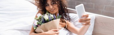 horizontal crop of curly woman taking selfie with flowers in bedroom clipart