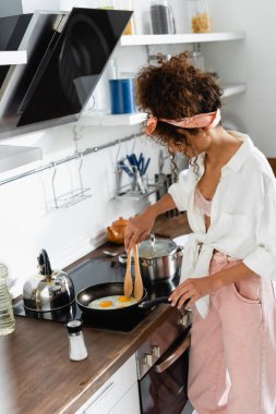 Mutfakta tavada yumurta pişirirken mutfak maşası tutan kıvırcık kadın. 