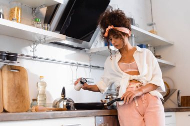 young curly woman holding bottle with salt above frying pan in kitchen  clipart