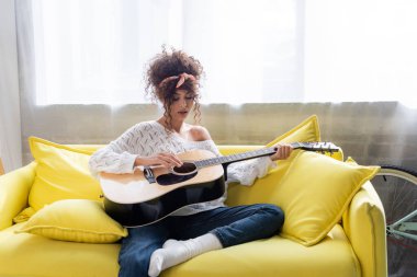 Oturma odasındaki kanepede akustik gitar çalan kıvırcık kadın. 