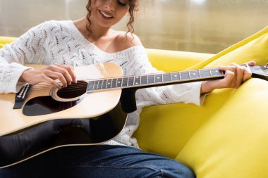 Oturma odasındaki kanepede akustik gitar çalan kıvırcık kadın görüntüsü. 