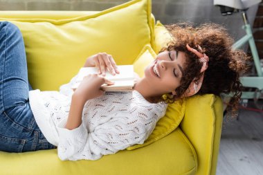 Oturma odasındaki kanepede uzanmış kitap tutan heyecanlı genç bir kadının yüksek açılı görüntüsü. 