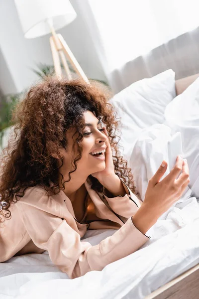 Excited Curly Woman Lying Bed Using Smartphone Bedroom — Stock Photo, Image