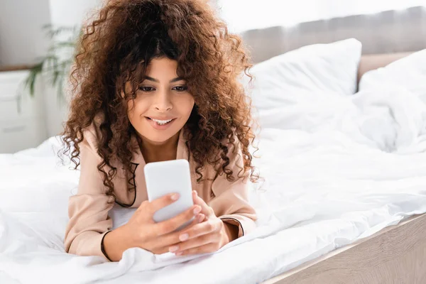 Young Curly Woman Biting Lip Using Smartphone Bedroom — Stock Photo, Image
