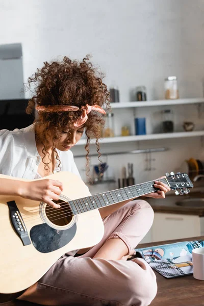 Joven Mujer Rizada Tocando Guitarra Acústica Cerca Taza Mesa — Foto de Stock