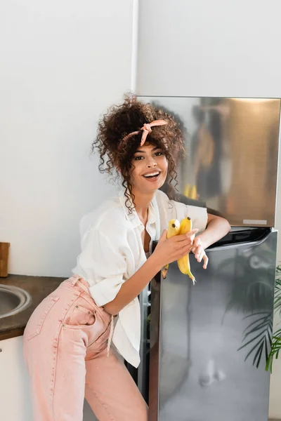 Радостная Женщина Держит Бананы Возле Холодильника Кухне — стоковое фото