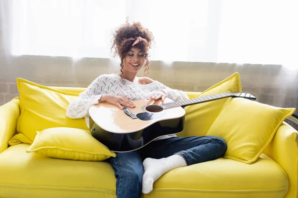 pleased woman holding acoustic guitar and sitting on sofa in living room