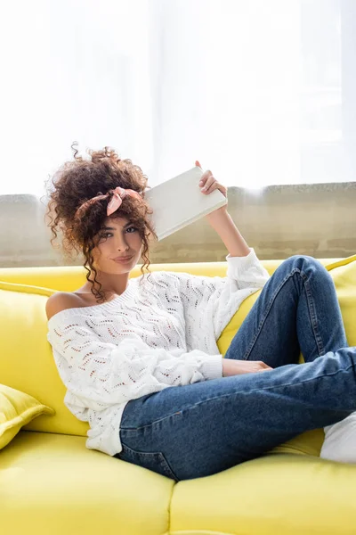 Kıvırcık Genç Bir Kadın Kitap Tutuyor Oturma Odasında Kameraya Bakıyor — Stok fotoğraf