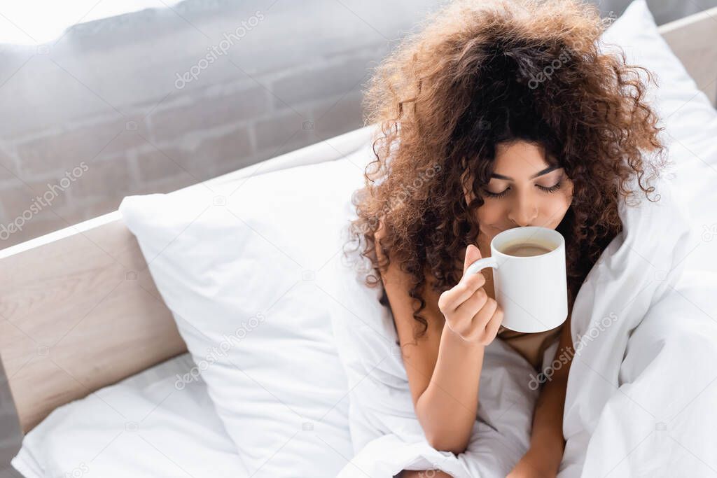 high angle view of young curly woman smelling coffee in bedroom