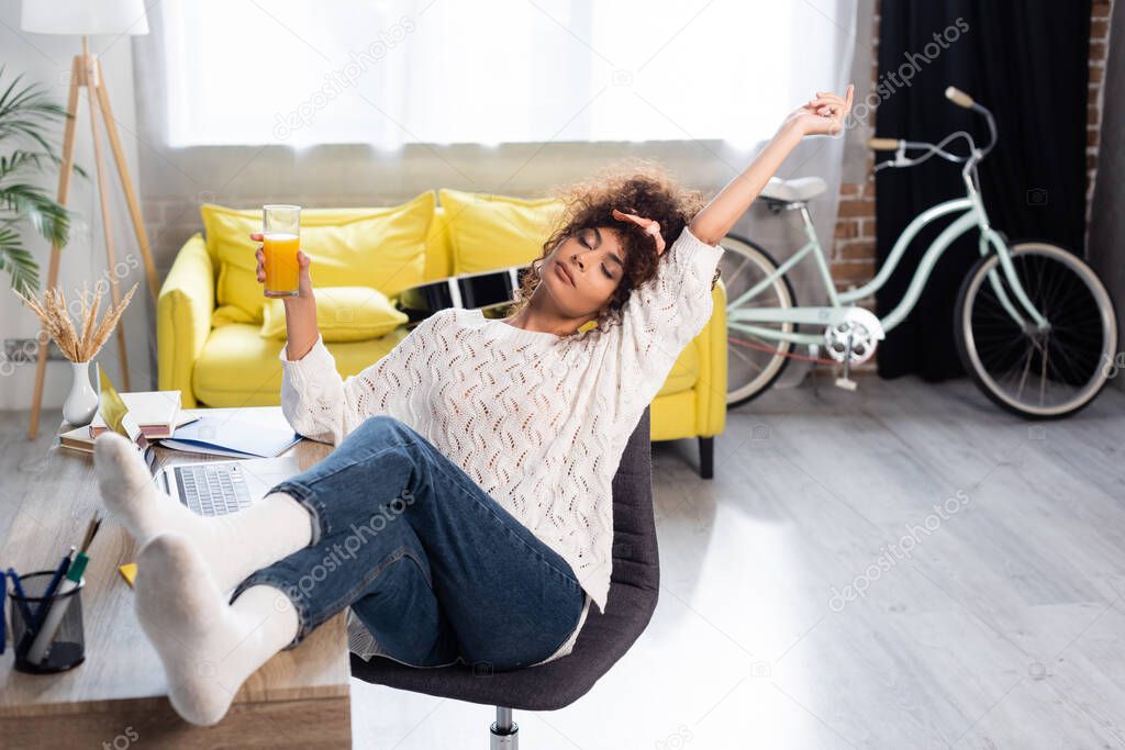 curly woman with closed eyes holding glass of orange juice while resting on chair near laptop