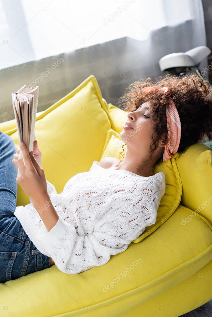 pleased young woman reading book while lying on sofa in living room 