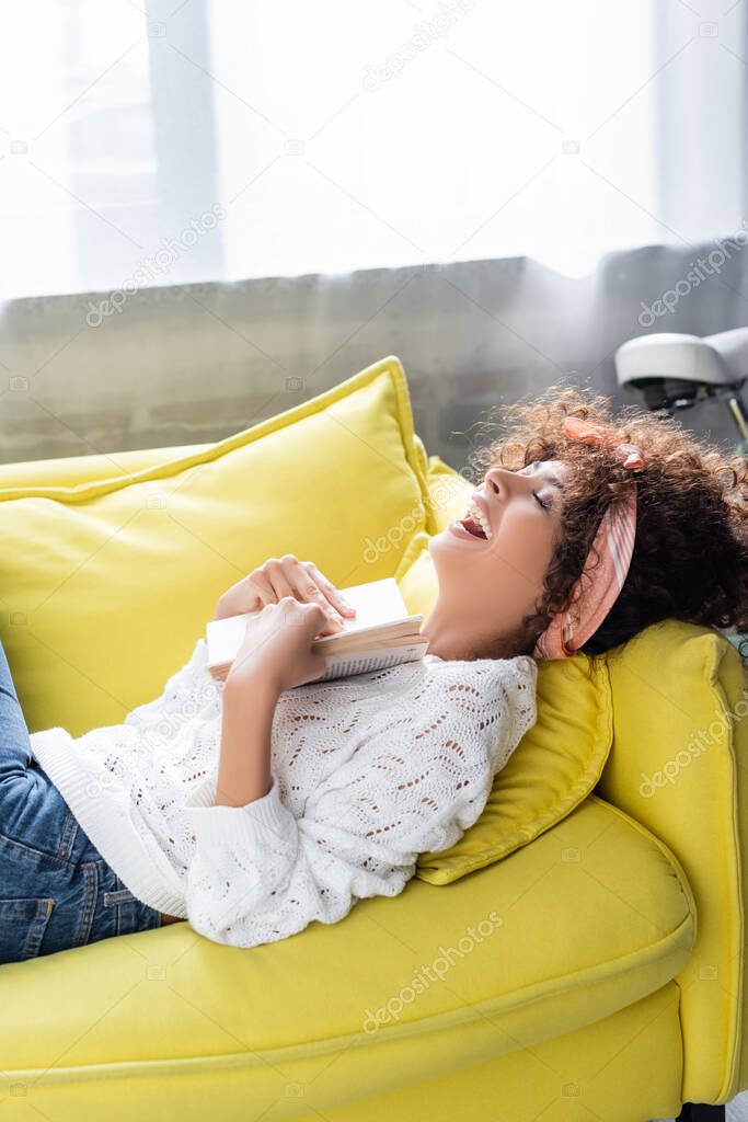 excited young woman holding book while lying on sofa in living room 
