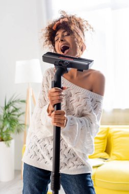 curly woman singing while holding vacuum cleaner at home clipart
