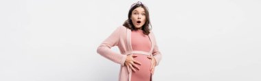panoramic shot of shocked and pregnant woman looking at camera while touching belly on white clipart