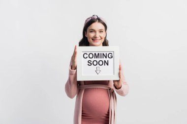 joyful pregnant woman holding board with coming soon lettering isolated on white clipart