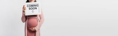 panoramic crop of pregnant woman holding board with coming soon lettering isolated on white clipart