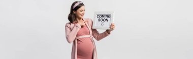 joyful pregnant woman pointing with finger at board with coming soon lettering isolated on white clipart
