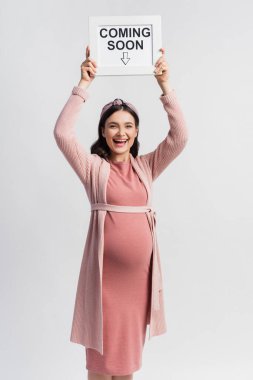 excited and pregnant woman holding board with coming soon lettering isolated on white clipart