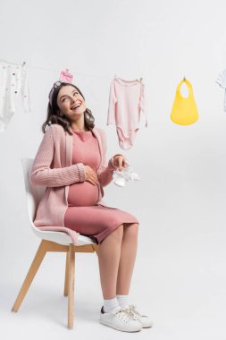 Hamile ve memnun bir kadın küçük çizmeler tutuyor ve bebek kıyafetlerinin yanında oturuyor.
