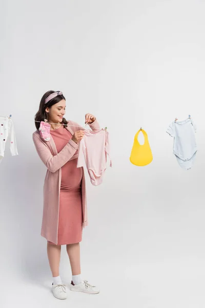 Neşeli Hamile Kadın Beyaz Elbise Üzerine Bebek Kıyafetleri Asıyor — Stok fotoğraf