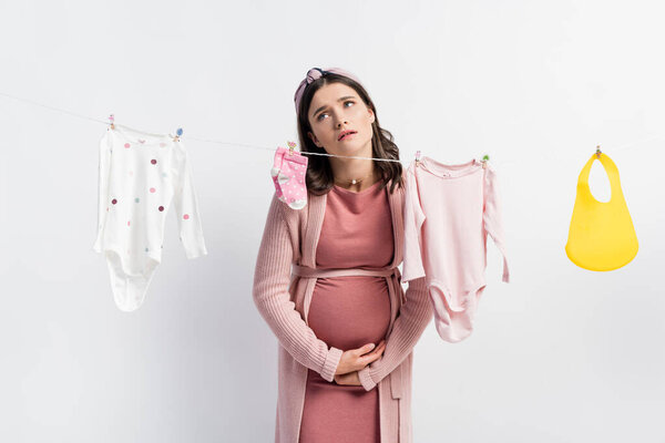 sad pregnant woman touching belly near baby clothes on clothing line isolated on white