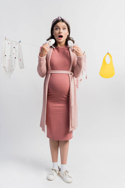 shocked and pregnant woman holding tiny boots near baby clothes on clothing line on white