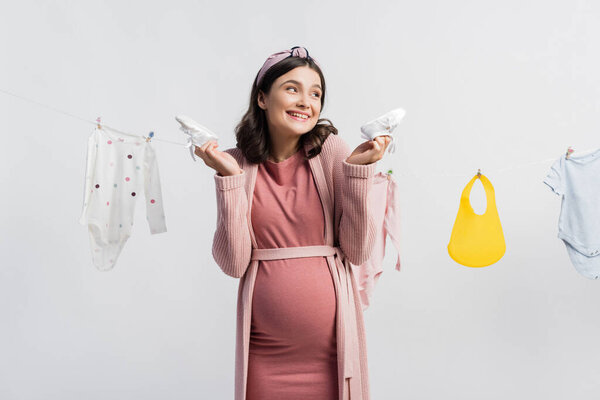 joyful pregnant woman holding tiny boots near baby clothes on clothing line isolated on white