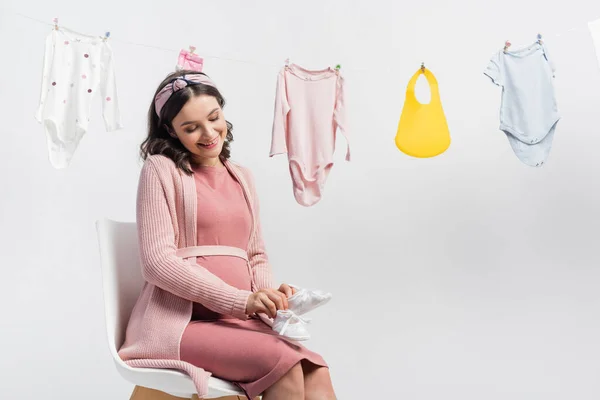 Mujer Alegre Embarazada Sosteniendo Pequeñas Botas Sentada Cerca Ropa Bebé — Foto de Stock