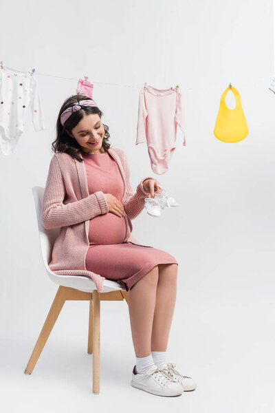 pleased pregnant woman holding tiny boots and sitting near near baby clothes on white