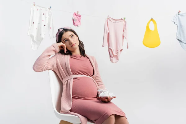 Zamyšlená Těhotná Žena Drží Malé Boty Sedí Blízkosti Dětské Oblečení — Stock fotografie