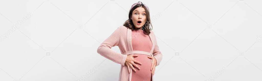 panoramic shot of shocked and pregnant woman looking at camera while touching belly on white